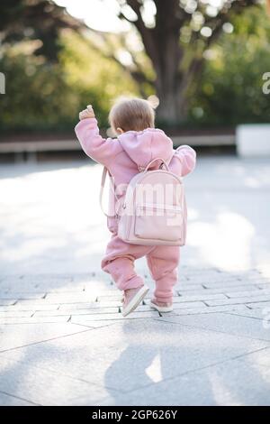 Divertente bambina 1 anno di età indossare tuta sportiva rosa elegante e zaino camminare sulla strada su sfondo urbano. Bambino piccolo carino con vestiti alla moda. CHIL Foto Stock