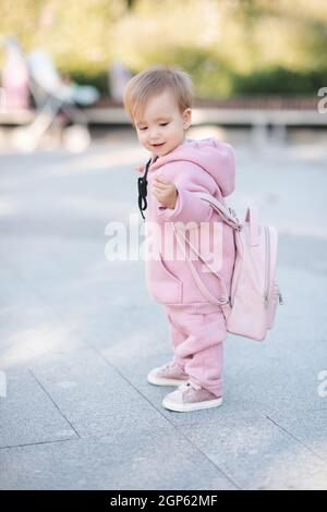 Simpatica ragazza divertente 1-2 anni indossare felpa rosa con cappuccio e zaino camminare in città strada oltre urbano all'aperto. Infanzia. Bambino piccolo m Foto Stock