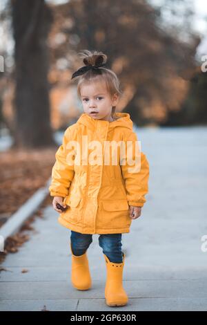 Bella ragazza divertente bambino 2-3 anni indossare giallo impermeabile luminoso, stivali di gomma a piedi nel parco sopra le foglie cadute all'aperto. Stagione autunnale. Felice bambino oltre Foto Stock