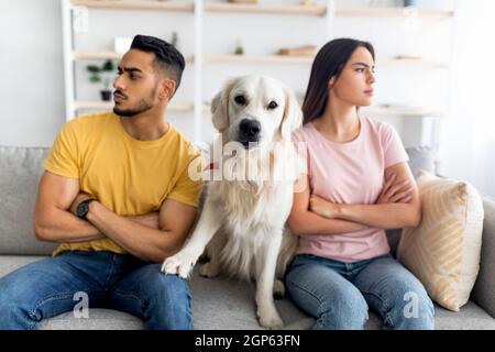 Sconvolto giovane coppia multinazionale che ha combattuto, guardando in direzioni opposte, carino cane seduto sul divano tra loro, in interni. Crisi di relazione, m Foto Stock
