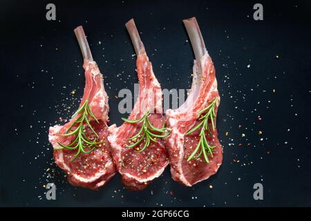 carne di agnello crudo in sella con osso sale, pepe decorato rosmarino vista dall'alto su sfondo nero Foto Stock