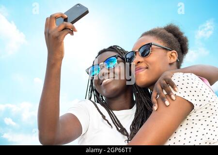 Primo piano ritratto di due diverse ragazze teen africane che indossano occhiali da sole prendendo auto ritratto con telefono contro il cielo blu. Foto Stock