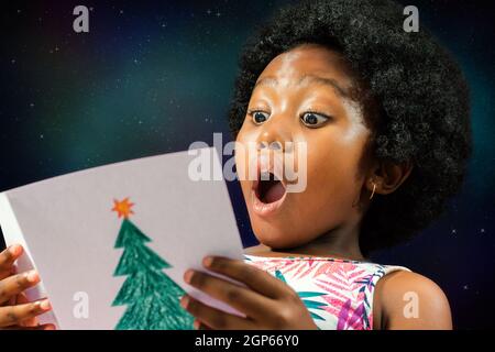 Primo piano faccia colpo di ragazza africana piccola lettura una scheda di natale con sorpresa volto Expression.Excited capretto con bocca aperta contro la stella galassia indietro Foto Stock