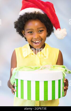 Primo piano ritratto di bambino africano felice che tiene natale scatole regalo.ragazza che indossa cappello rosso su sfondo azzurro chiaro. Foto Stock