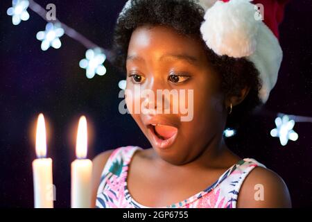 Primo piano in faccia colpo di uscita piccola ragazza africana guardando candele incandescente nel buio. Ritratto basso dell'ascensore del bambino che indossa il cappello di natale contro la stella Foto Stock