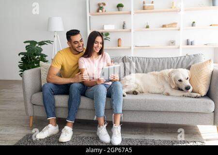 Bella giovane coppia diversificata seduta sul divano con il loro cane carino, utilizzando il PC tablet, navigare sul web a casa, spazio copia. Coniugi multirazziali millenari Foto Stock