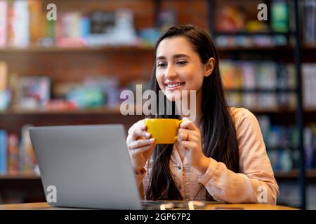 Donna araba millenaria che si diverte a bere un caffè mentre lavora su un computer portatile in un bar, sorridendo e guardando lo schermo del pc. Giovane freelance con bre Foto Stock