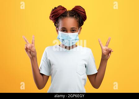 Carina ragazza afroamericana teen in maschera protettiva viso mostrando gesto di pace con entrambe le mani, indossare maschera medica mentre COVID-19 blocco, in posa su Foto Stock