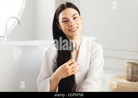 Bella ragazza con massaggio drenante rullo di faccia seduta in bagno di lusso. Foto Stock