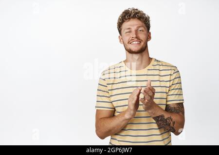 Bell'uomo biondo riccio-pelo che stringe le mani e sorride contento, complimentandosi, applauso, in piedi su sfondo bianco Foto Stock