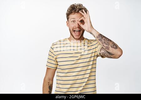 L'uomo biondo felice guarda attraverso il segno di zero buono, mostra il gesto OK e sorridendo stupito, reazione alla vendita, promozione negozio, in piedi su sfondo bianco Foto Stock