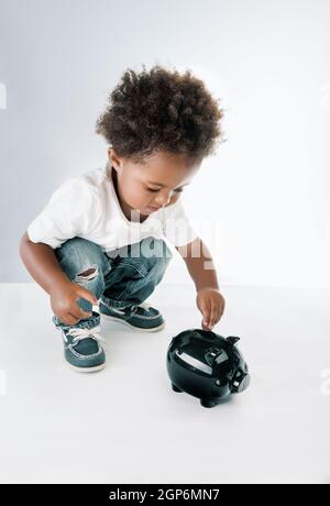 Ritratto di un bel bambino afro-americano che mette le monete nella scatola di denaro a forma di maiale. Isolato su sfondo bianco grigio. Speranza di un futuro migliore Foto Stock
