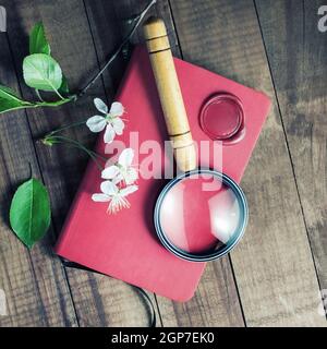 Taccuino rosso, lente d'ingrandimento, sigillo di cera, fiori di herry e foglie verdi su sfondo tavola di legno vintage. Foto Stock