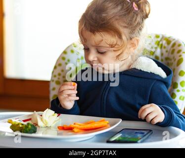 Il Toddler mangia nel seggiolone mentre guardi un film sul telefono cellulare. Molti bambini che non vogliono mangiare se non guardare il dispositivo mobile o t Foto Stock