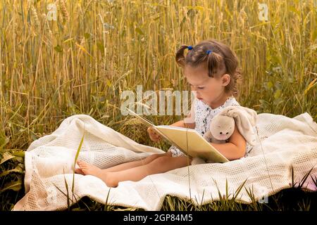 Piccola ragazza carina 1-3 in un vestito leggero abbraccia una lepre di peluche e legge un libro, seduto su un plaid in un campo di spikelets di segale in estate. Foto Stock