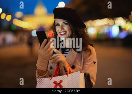 Ciao autunno. Donna elegante sorridente in trench beige con borse per lo shopping invio di messaggi di testo con smartphone fuori sulla strada della città in autunno Foto Stock