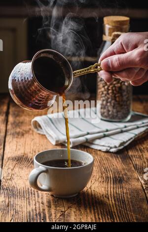 Versare il caffè turco in una tazza bianca. Vasetto di chicchi di caffè tostati su sfondo Foto Stock