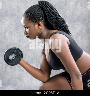 Primo piano ritratto di ragazza teen africana che si sta allenando con dumbbell.Side vista della giovane donna in abbigliamento sportivo su sfondo grigio. Foto Stock