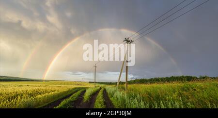 Vista panoramica sullo splendido paesaggio russo. Doppio arcobaleno su campi di grano verde, linee elettriche e strada di campagna Foto Stock