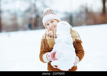 felice ragazza moderna in un cappello lavorato a maglia e cappotto in pelle di pecora con mittens e pupazzo di neve fuori nel parco della città in inverno. Foto Stock