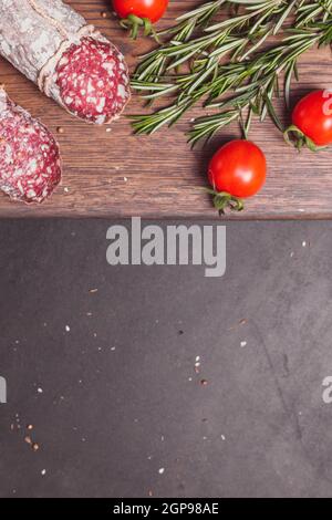 Salsiccia di prosciutto affumicata e fette tagliate con rosmarino e pomodori ciliegini su tavola di legno scuro. Vista dall'alto, layout piatto Foto Stock