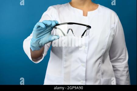 medico femminile in un abito medico bianco si trova su uno sfondo blu e contiene occhiali di plastica protettiva, primo piano Foto Stock