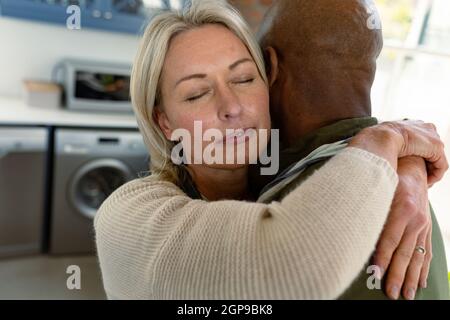 Felice senior diverse coppia in cucina indossando grembiuli, abbracciando Foto Stock