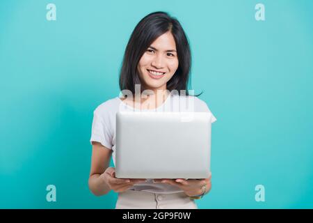 Ritratto felice asiatico bella giovane donna sorriso bianco denti in piedi indossare t-shirt bianca, lei tenendo e utilizzando un computer portatile, studio girato su blu Foto Stock