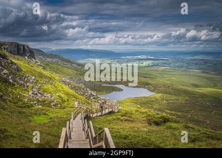 Persone che camminano su ripide scale di legno passerella nel Parco Monte Cuilcagh. Vista sul lago e sulla valle sottostante con il cielo spettacolare, Irlanda del Nord Foto Stock