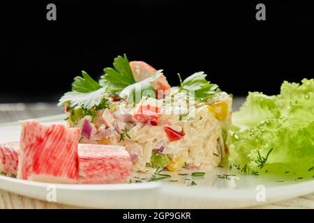 Insalata appetitosa da bastoncini di granchio con riso e verdure su piatto bianco adagiato su tavola di legno su sfondo nero. Foto Stock