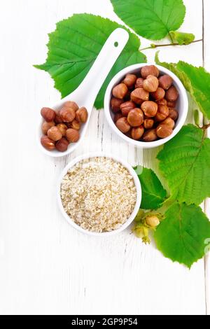 Farina e nocciole in due ciotole, un cucchiaio con noccioli di noce pelati e un ramo di filbert con foglie verdi su fondo di legno dall'alto Foto Stock