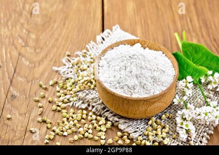 Farina di grano saraceno da cereali verdi in una ciotola su insaccatura, semole sulla tavola, fiori freschi e foglie di grano saraceno sullo sfondo di una b di legno marrone Foto Stock