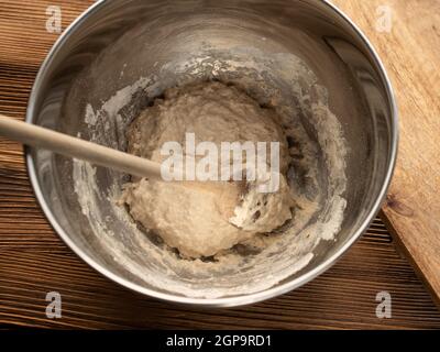 Impastare in un recipiente di metallo per una baguette fatta in casa. Mescolando gli ingredienti con un cucchiaio di legno su un banco da cucina in legno. Foto dall'alto. Foto Stock