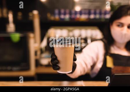 Il barista in maschera protettiva e guanti neri offre una tazza di caffè al bar. Foto di alta qualità Foto Stock