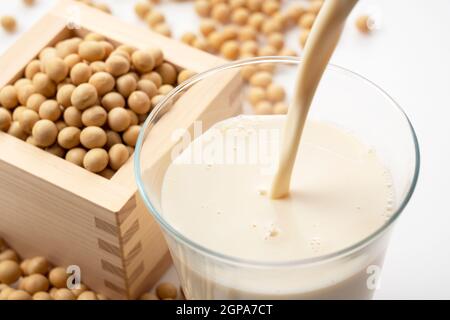 Versare il latte di soia in un bicchiere posto su uno sfondo bianco. Fagioli di soia in un Masu sullo sfondo Foto Stock