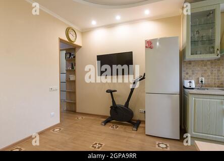 Un frammento dell'interno del soggiorno combinato con la cucina, una TV appeso alla parete, una cyclette e un frigorifero sono situati nelle vicinanze Foto Stock