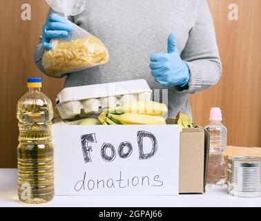 donna in un maglione grigio mette in una scatola di cartone vari alimenti, frutta, pasta, olio di girasole in una bottiglia di plastica e confetture. Donazione e volontariato Foto Stock