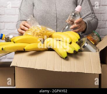 donna in un maglione grigio mette in una scatola di cartone vari alimenti, frutta, pasta, olio di girasole in una bottiglia di plastica e confetture. Donazione e volontariato Foto Stock