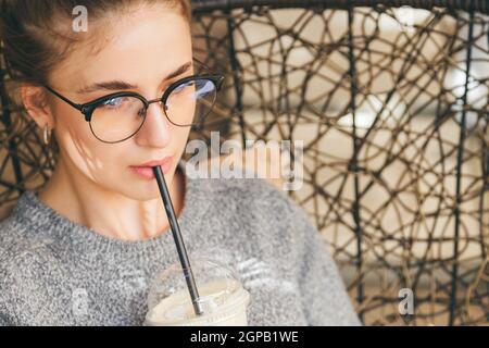 Primo piano ritratto di giovane milleniale hipster donna drink cocktail in un bar all'aperto. Casual vestito con occhiali. Foto Stock