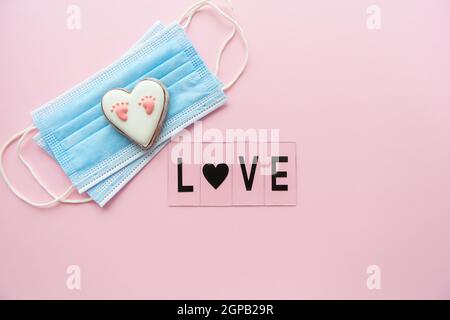 Maschera medica monouso, cuore con impronta del bambino su sfondo rosa. Parto durante la pandemia del coronavirus. Lettere d'amore Foto Stock