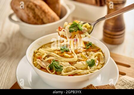 Zuppa di noodle. Deliziosa zuppa con noodle, pezzi di pollo e verdure in ciotola bianca servita con spezie e pane su sfondo di legno chiaro. Foto Stock