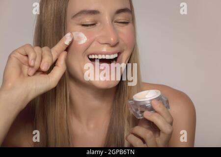 Ritratto di una bella ragazza sorridente Laughing e applicazione crema idratante viso isolato su sfondo Beige. Cosmetici naturali. Stile di vita sano. BEA Foto Stock