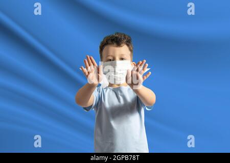 Piccolo ragazzo bianco in una maschera protettiva sullo sfondo della bandiera della Somalia. Fa un segno di stop con le mani, stare a casa Somalia. Foto Stock