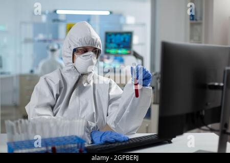 Donna ricercatrice biologa che tiene in mano la provetta per l'analisi delle competenze in materia di liquidi lavorando allo sviluppo del vaccino del coronavirus in laboratorio. Scienziato che indossa il vestito ppe durante l'esperimento medico Foto Stock