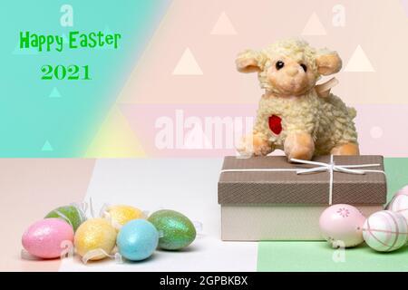 Biglietto d'auguri di Pasqua. Primo piano di uova colorate, una confezione regalo con un agnello pasquale su un tavolo multicolore di fronte a sfondo astratto pastello wit Foto Stock