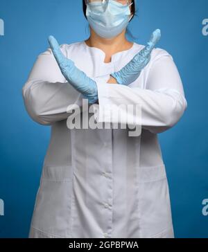 medica femminile in un cappotto bianco, una maschera si erge su uno sfondo blu, braccia incrociate, un gesto sufficiente Foto Stock