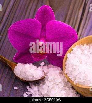 Il sale da bagno bianco in una tazza di legno con un cucchiaio su un tavolo di legno con un'orchidea Foto Stock