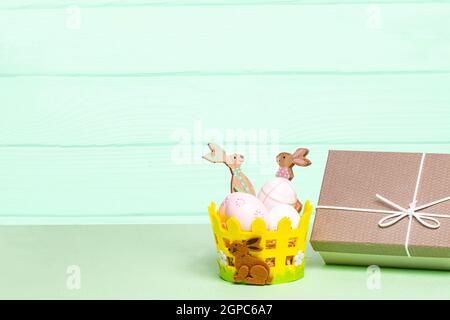 Modello del biglietto di auguri di buona Pasqua. Composizione decorativa del cesto con uova colorate e una scatola regalo su un tavolo verde su sfondo verde chiaro. Foto Stock