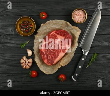 Primo piano una bistecca di manzo crudo marmorizzato invecchiato su carta marrone pergamena che avvolge, con coltello e spezie, su sfondo di legno nero, el Foto Stock