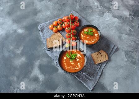 Zuppa invernale calda. Zuppa di pomodoro rosso caldo con aglio, paprika dolce, prezzemolo, servito con panna e pane di pasta madre in due ciotole di ceramica nera su una b grigia Foto Stock